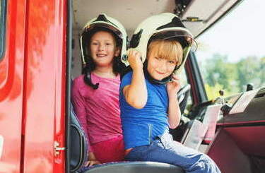 Dwoje dzieci siedzi w kaskach w samochodzi strażackim, widok przed boczne drzwi.