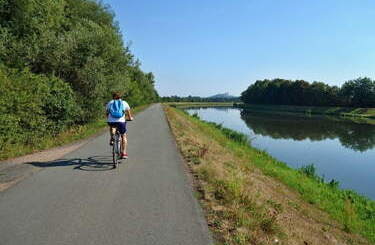 Ścieżka bieglnąca wzdłuż rzeki, a na jedzie niej rowerzysta w kasku.